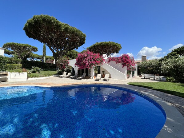 A swimming pool with a white structure and pink flowering plants nearby.