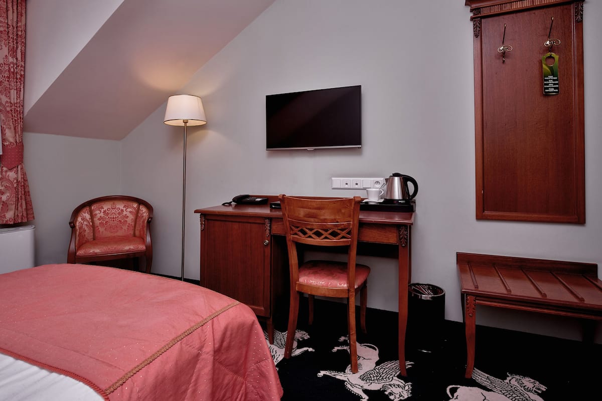 A hotel room with a bed, a desk with a chair, a television, a wardrobe, and a floor lamp.