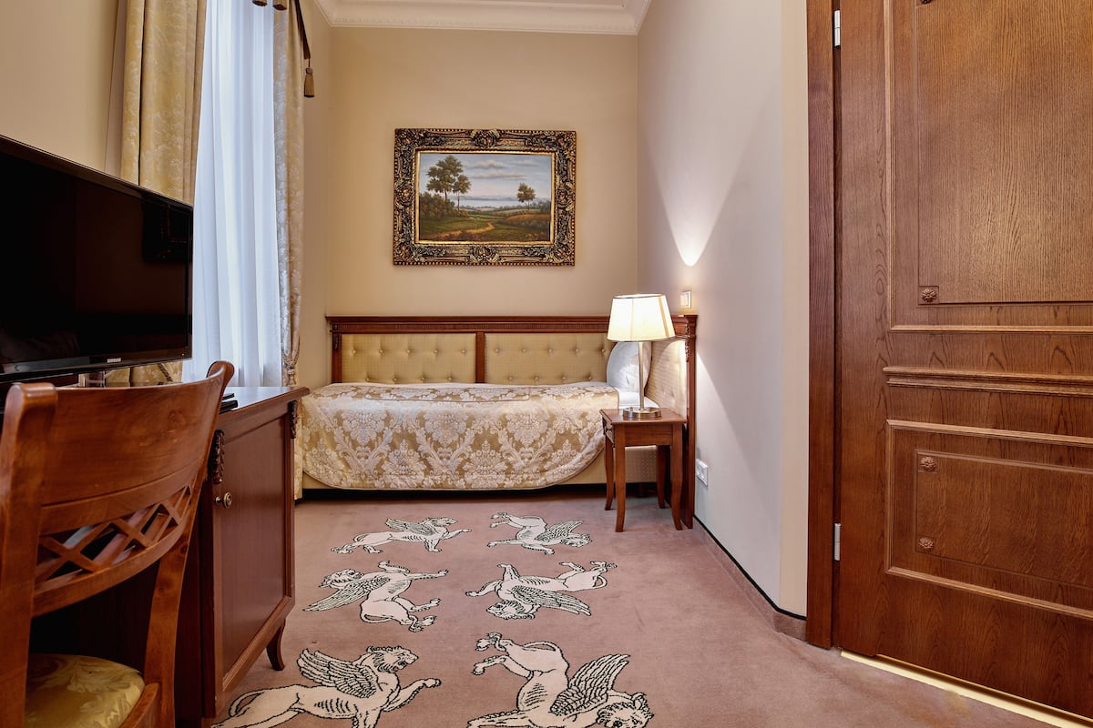 A hotel room with a bed, a television, a nightstand with a lamp, a wooden wardrobe, and a carpet with a bird design.