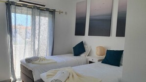 Habitación de hotel con dos camas, una ventana con cortinas traslúcidas, aire acondicionado de pared y una mesita de noche con lámpara.