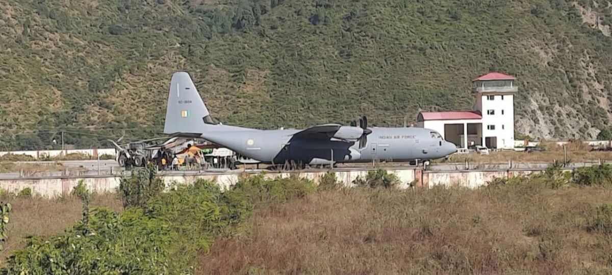 Un grand avion militaire est stationné sur une piste d’atterrissage, avec une chaîne de montagnes en arrière-plan.