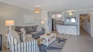 A modern living room with a grey sofa, striped armchairs, and a white coffee table. A kitchen with white appliances and bar stools is visible in the background.