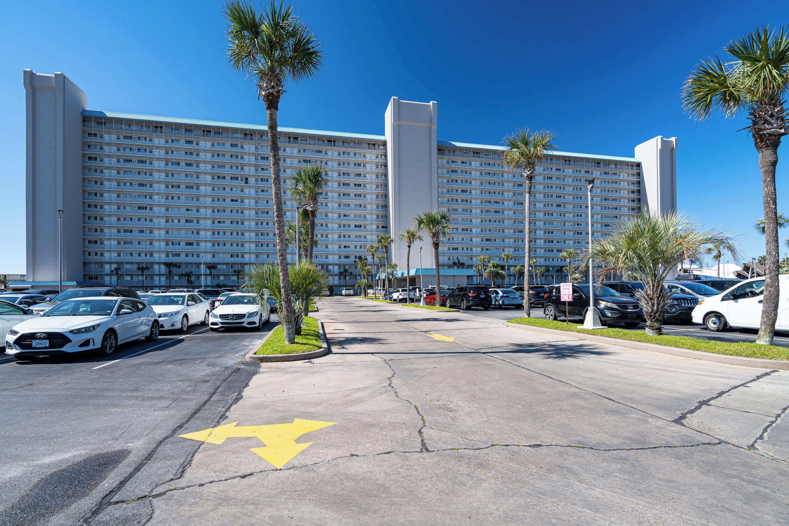 Un grande hotel con diversi piani, un parcheggio con numerose auto e palme.