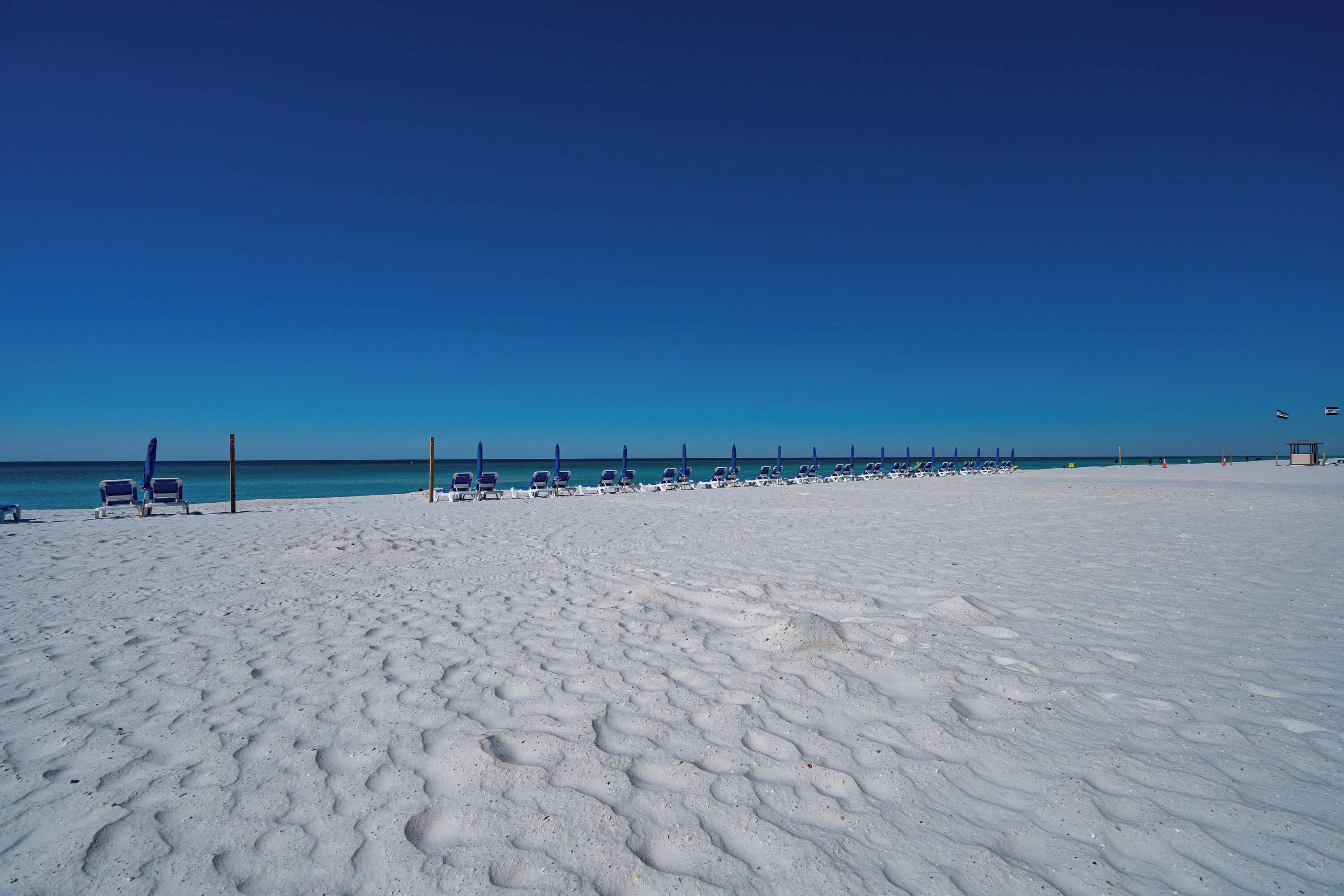 A beach with rows of sun loungers and umbrellas, a clear blue sky, and the ocean in the background.