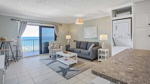 A living room with a grey sofa, a coffee table, a dining area with a table and chairs, a kitchen with a stove, and a view of the sea through a large window.