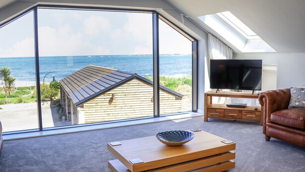 Ein großzügiges Wohnzimmer mit einem großen Erkerfenster mit Meerblick, einem Holztisch, einer Ledersofa und einem Flachbildfernseher.