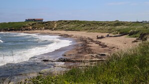 Ein Strand mit einem Haus im Hintergrund, grasbewachsenen Dünen und einer steinigen Küstenlinie.