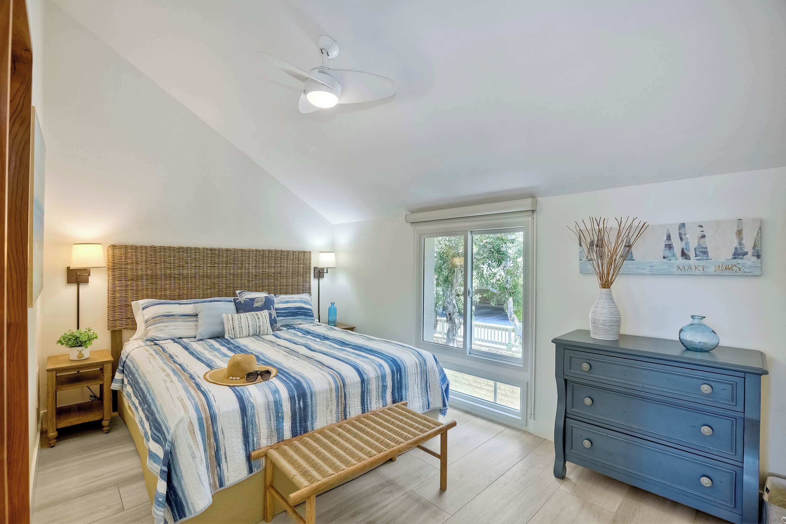 A bedroom with a bed, bedside table, lamp, bench, dresser, and a window with a view.