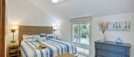 A bedroom with a bed, bedside table, lamp, bench, dresser, and a window with a view.