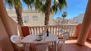 Une terrasse avec une table ronde dressée pour quatre personnes, entourée de chaises blanches, de palmiers et offrant une vue sur les bâtiments.