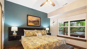 A bedroom with a large bed, a nightstand with a lamp, a bench, and a window with a view of trees. - That Lakefront Taneycomo House - Branson (Branson)