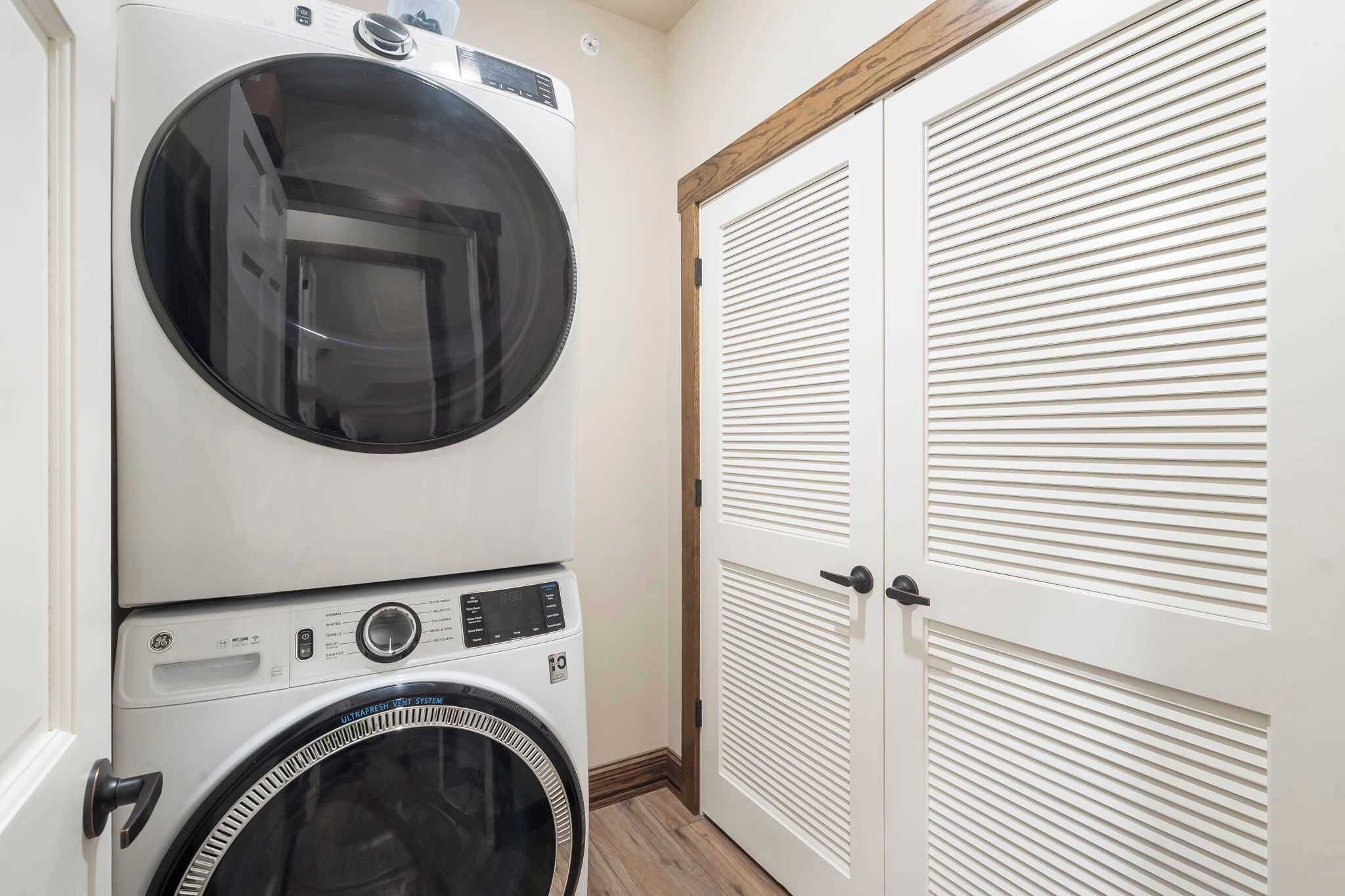 Un cuarto de lavandería con lavadoras y secadoras apiladas, una puerta de madera y un armario blanco con puertas persianas.