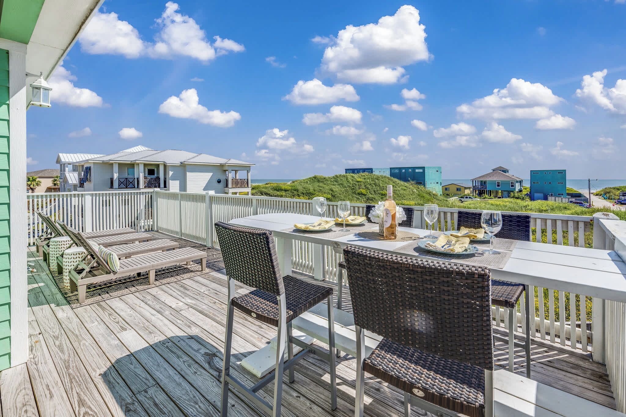 A wooden deck with outdoor furniture, a table set for dining, and a view of the ocean and buildings in the background.