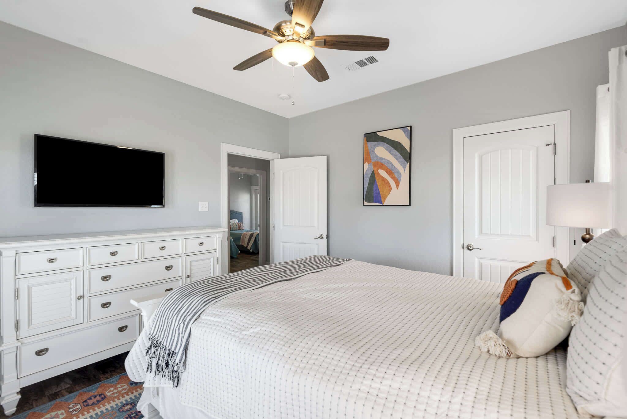 Une chambre à coucher avec un lit, un ventilateur de plafond, une télévision, une commode et une porte.