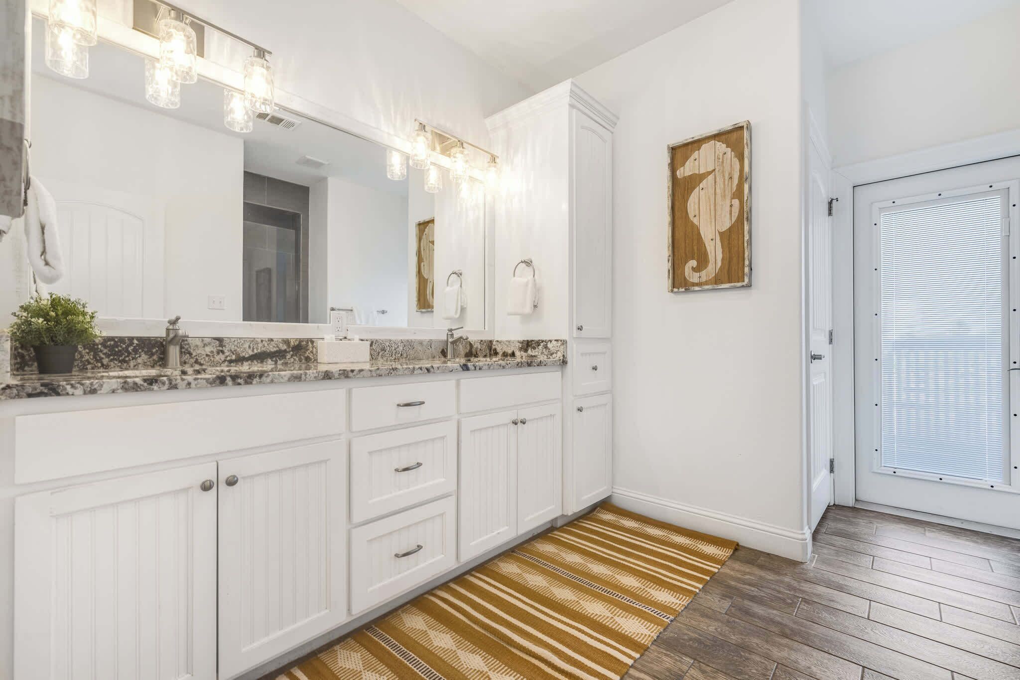 A bathroom with a large mirror, white cabinets, a granite countertop, and a patterned rug.