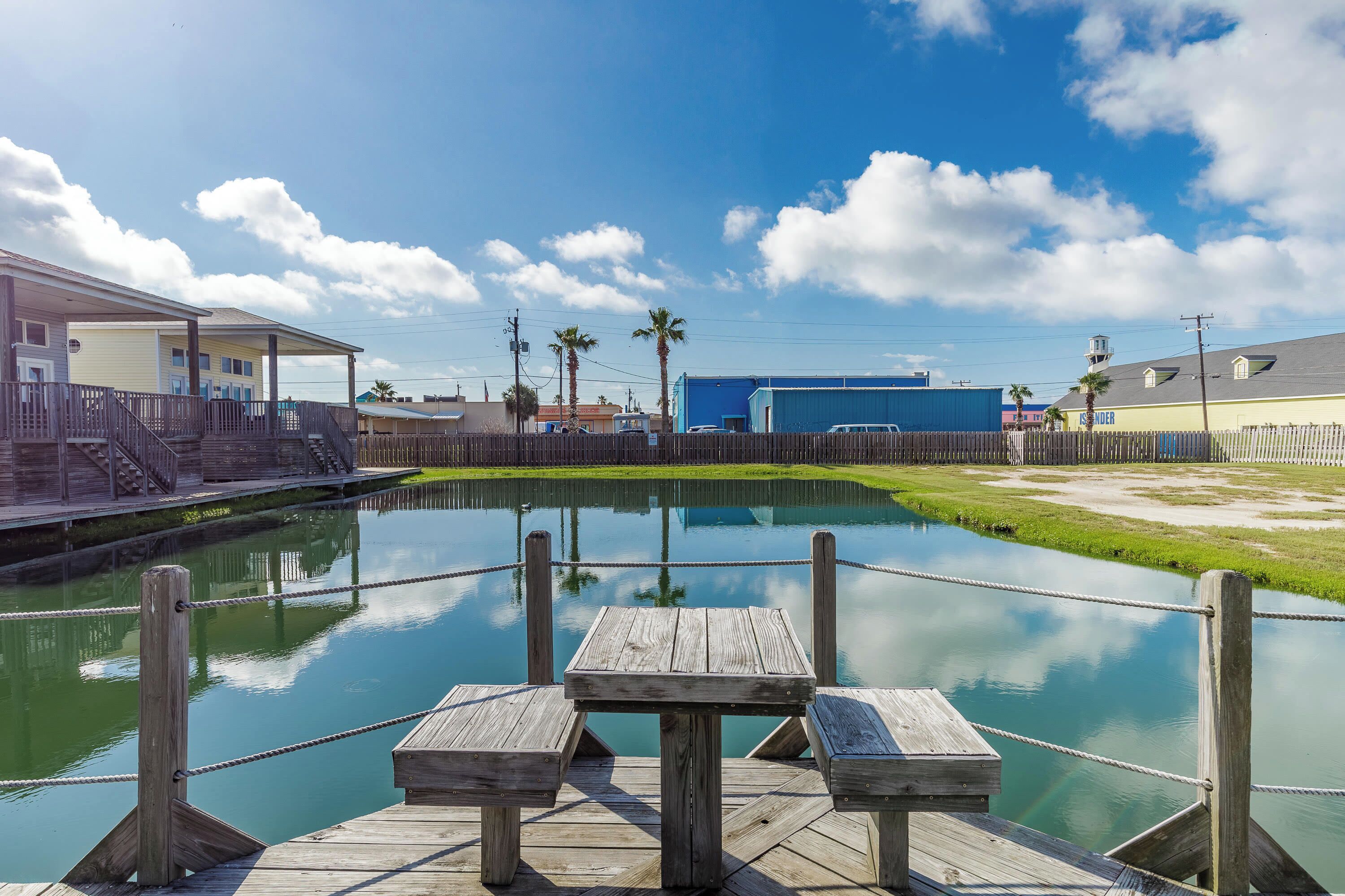 A wooden dock with a table and benches overlooking a calm water body, surrounded by buildings and a clear sky.