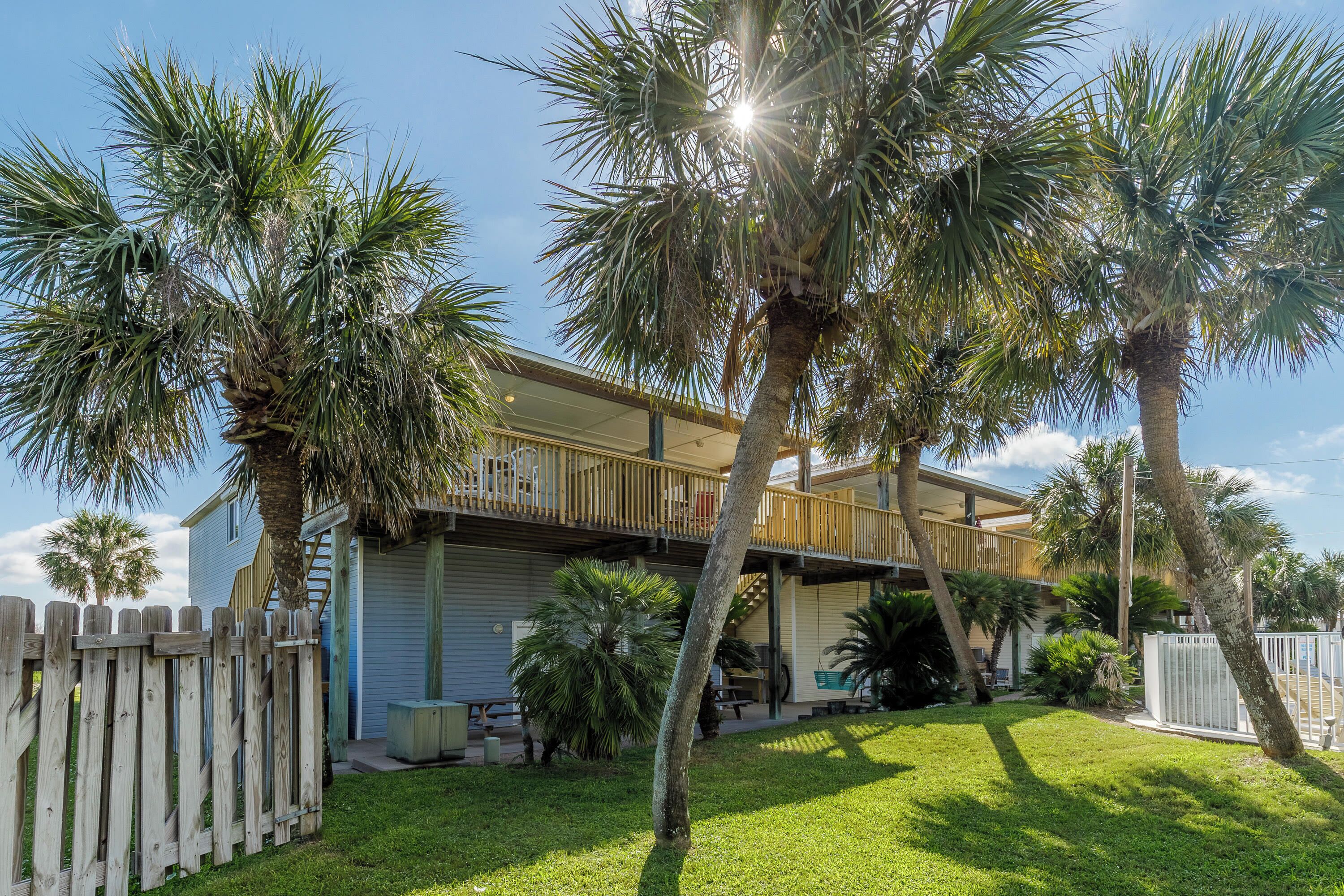 A two-story house with a balcony, surrounded by palm trees and a wooden fence.