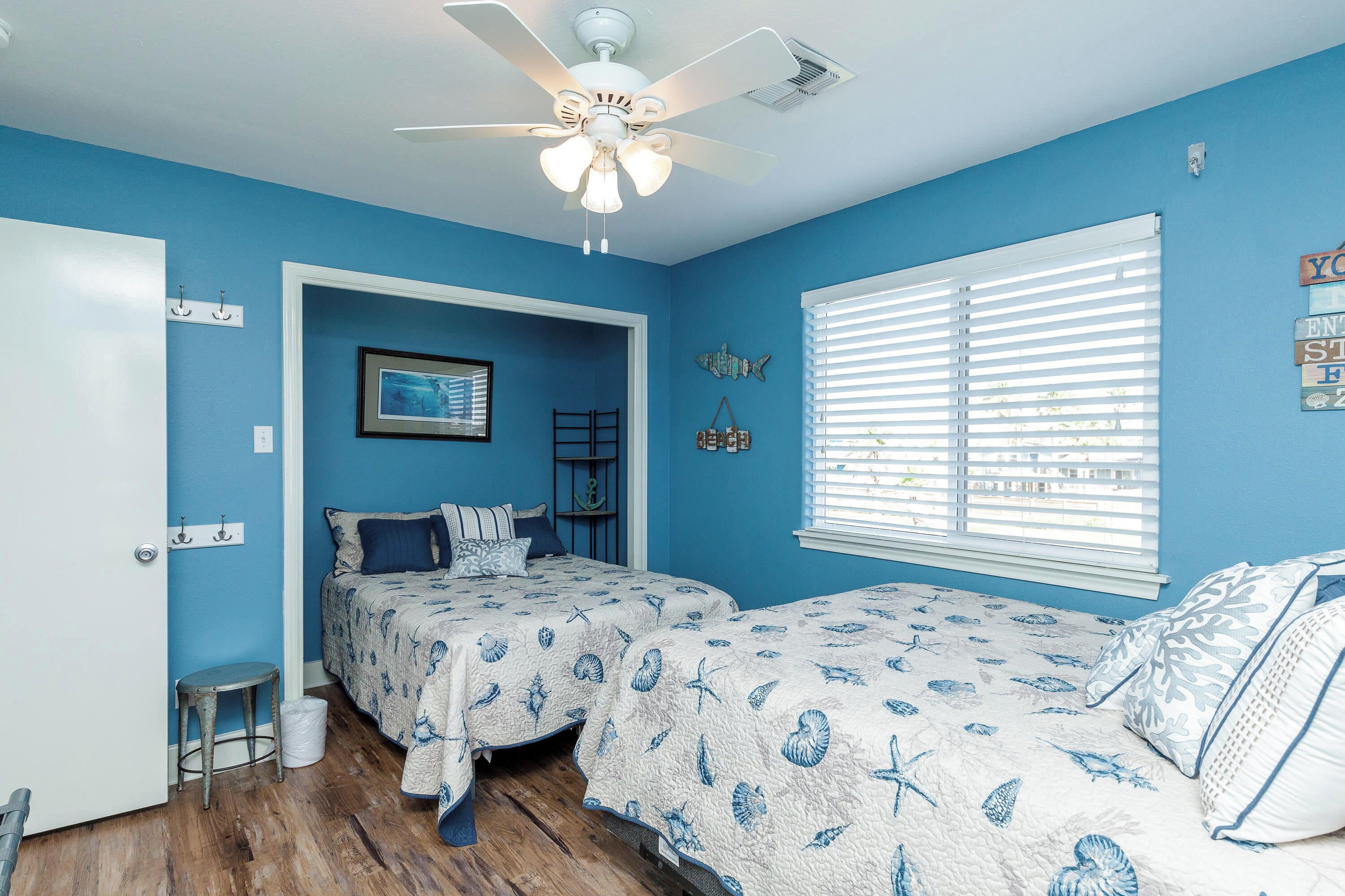 A bedroom with two beds, a ceiling fan, a window with blinds, and a door.