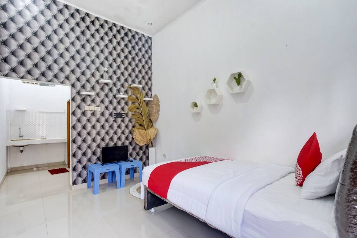 Un dormitorio moderno con una tapicería capitonada en la cabecera, una cama con ropa de cama blanca y roja, un taburete azul, un televisor sobre un soporte y estantes empotrados con plantas.