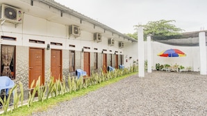 A building with multiple rooms, each with a door and an air conditioning unit. There is a gravel path leading to a covered area with a colorful umbrella and potted plants. - OYO 90124 Oemah Ratu Hostel Syariah (Bandar Lampung)