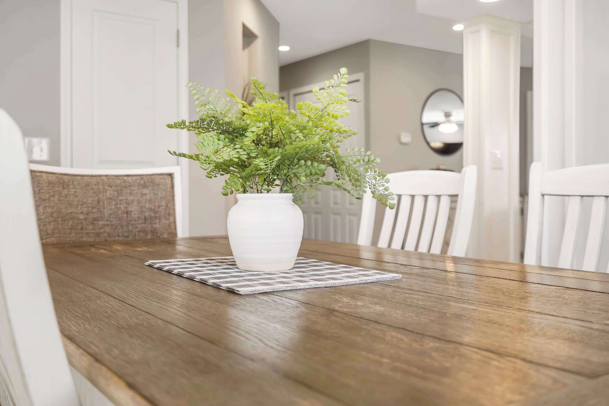 A dining table with a white vase containing green foliage, a checkered placemat, and a wooden chair.