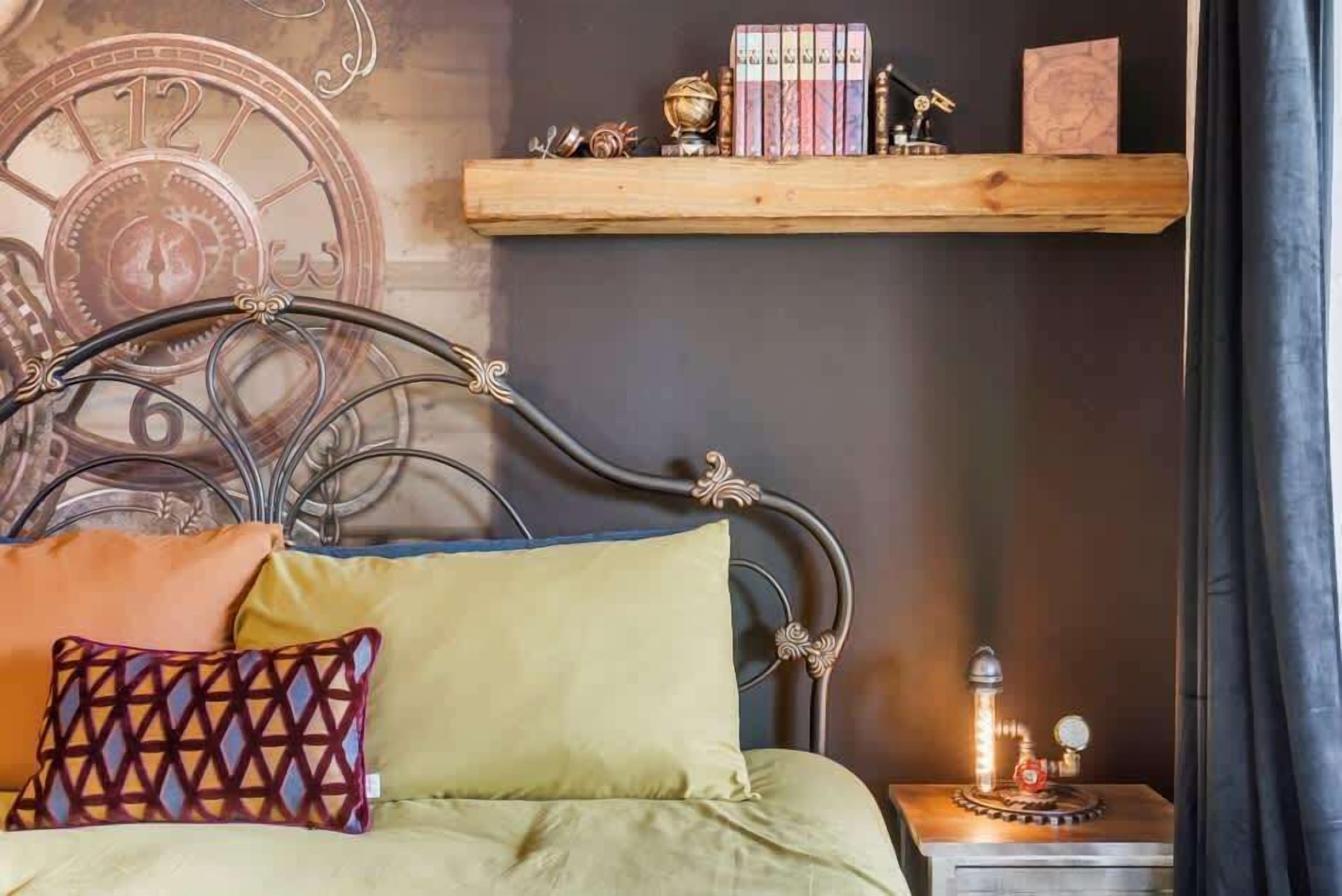 Uma cama com cabeceira de metal, uma mesa de cabeceira com abajur e uma estante com livros e objetos decorativos.