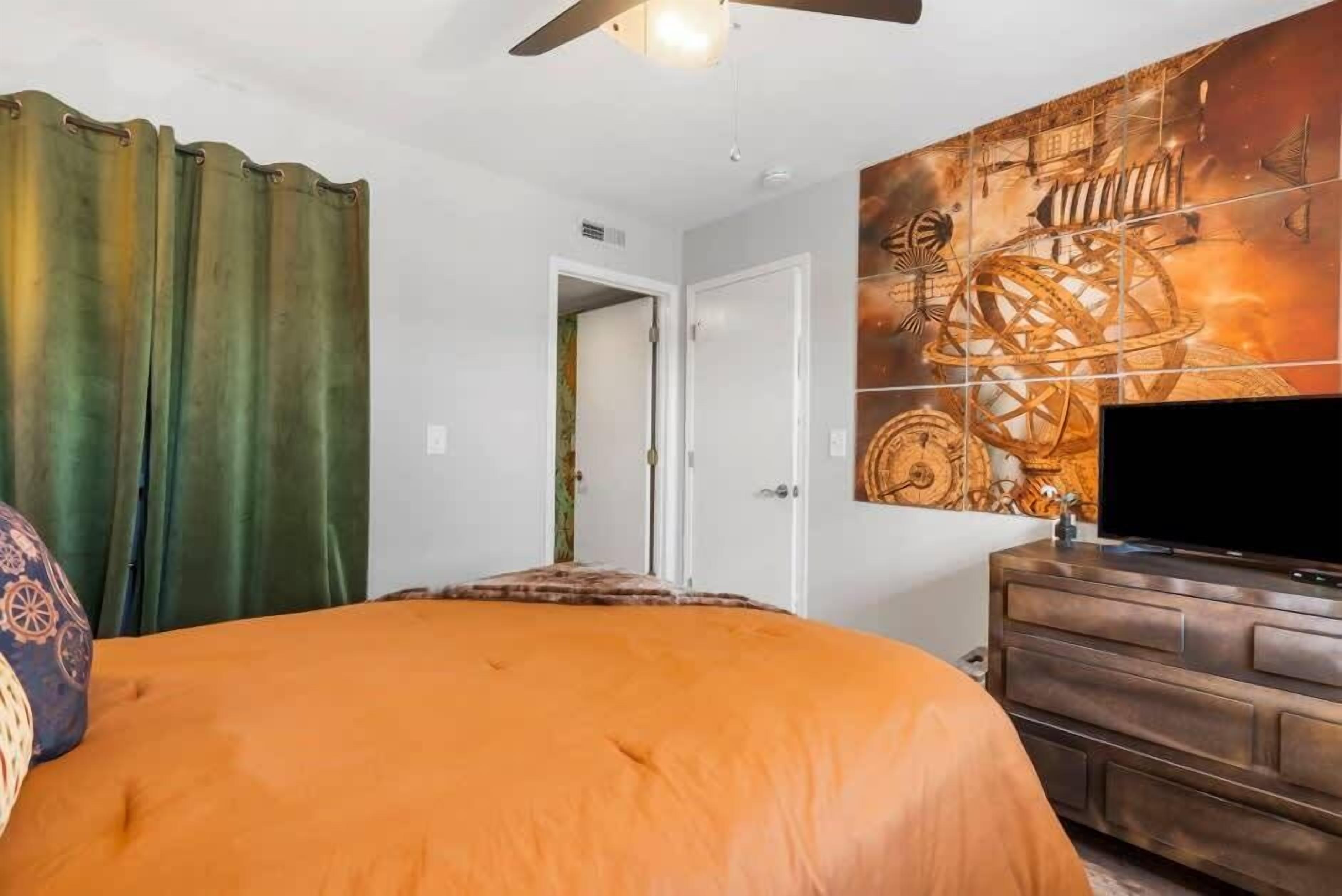 A bedroom with a bed, curtains, a dresser, a television, and a door leading to another room.