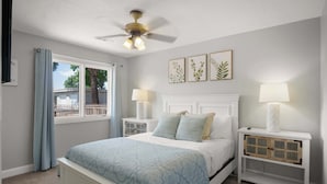 Un dormitorio con cama, dos mesitas de noche, ventilador de techo, ventana con cortinas y una obra de arte colgada en la pared.