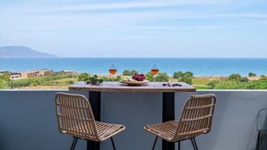 A balcony with two wicker chairs, a table with a plate of grapes and two glasses of red wine, and a view of the sea and distant buildings. - Blue Harmony villa SEA VIEW heated pool  (ΑΠΟΚΟΡΩΝΑΣ ΧΑΝΙΩΝ)