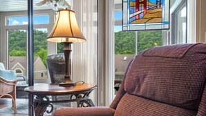 Une pièce chaleureuse avec un fauteuil en osier, une table avec une lampe et une vue sur la verdure grâce à de grandes fenêtres.
