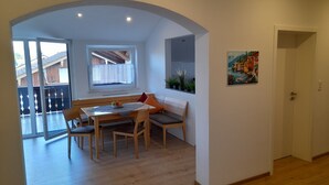 Een eetgedeelte met een houten tafel en stoelen, een bank met kussens, een raam met uitzicht en een deur naar een andere kamer.