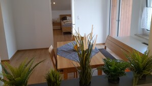 Een eethoek met een houten tafel en stoelen, omringd door potplanten. Een bed is zichtbaar op de achtergrond.