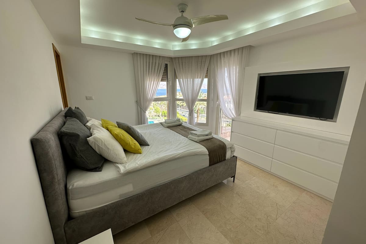 A modern bedroom with a bed, curtains, a ceiling fan, and a TV mounted on the wall.