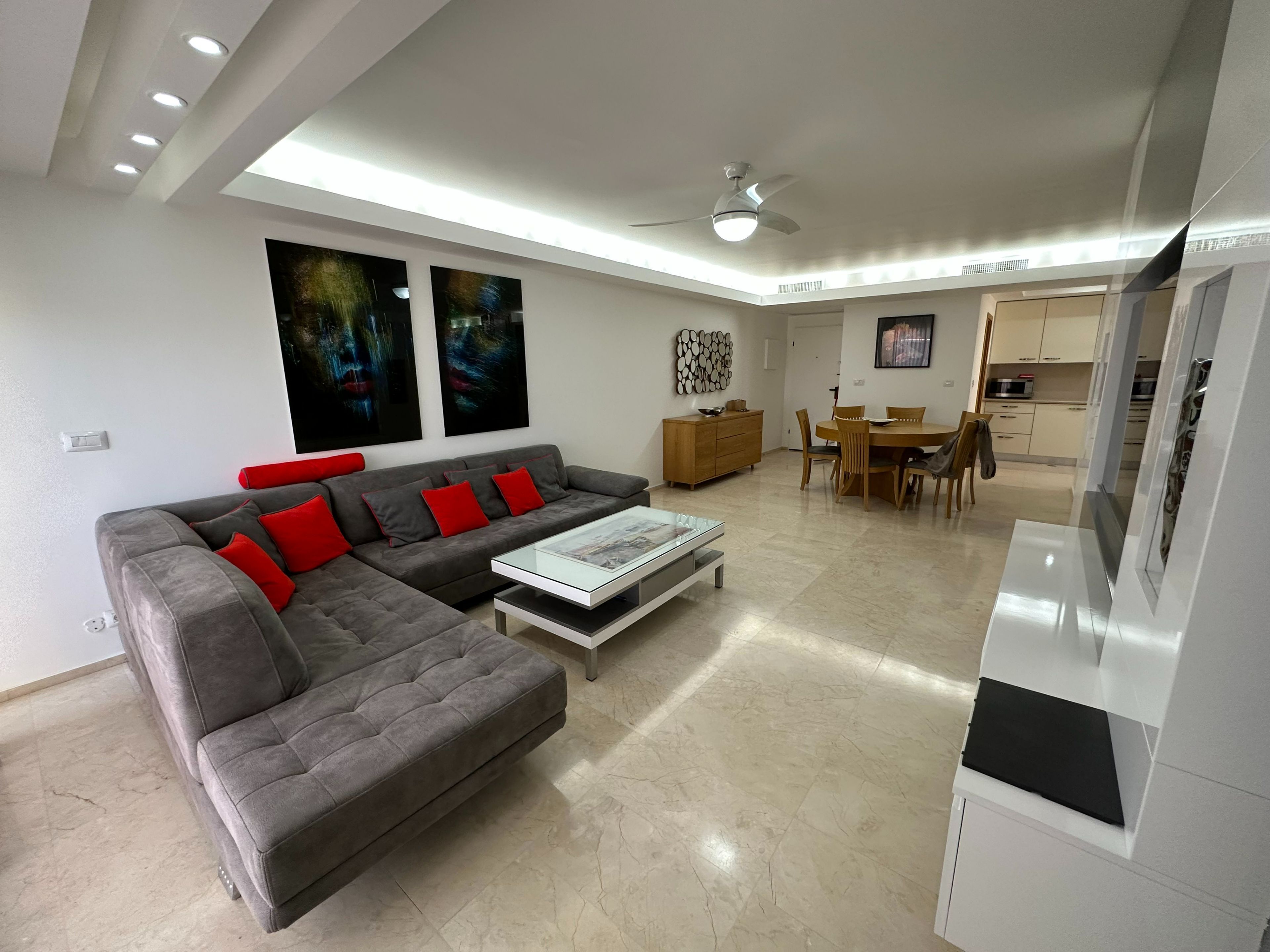 A modern living room with a grey sectional sofa, a glass coffee table, and a dining area with wooden furniture.