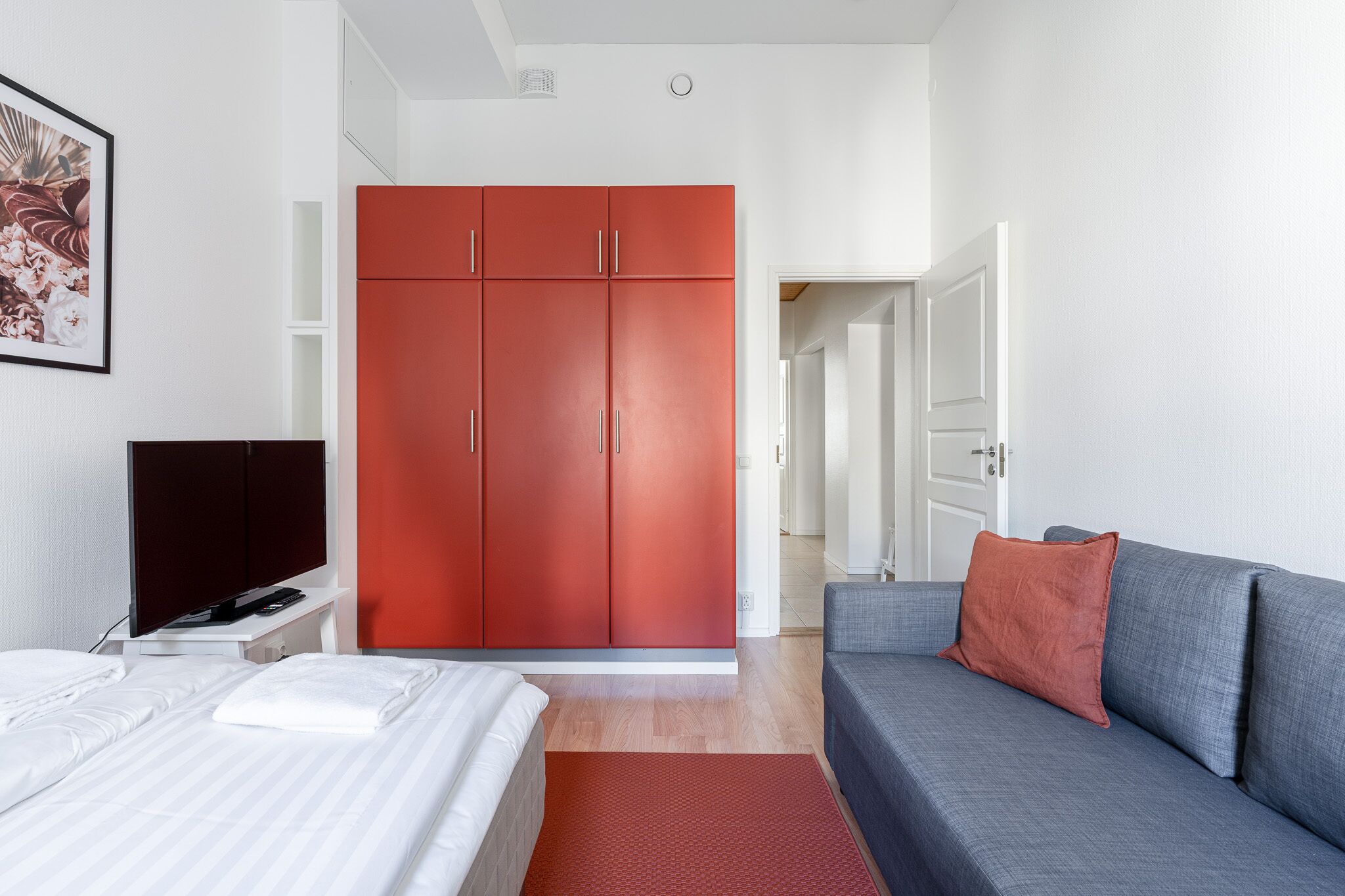 Et moderne hotelværelse med en seng, en grå sofa, et rødt klædeskab og et fjernsyn på et hvidt natbord.