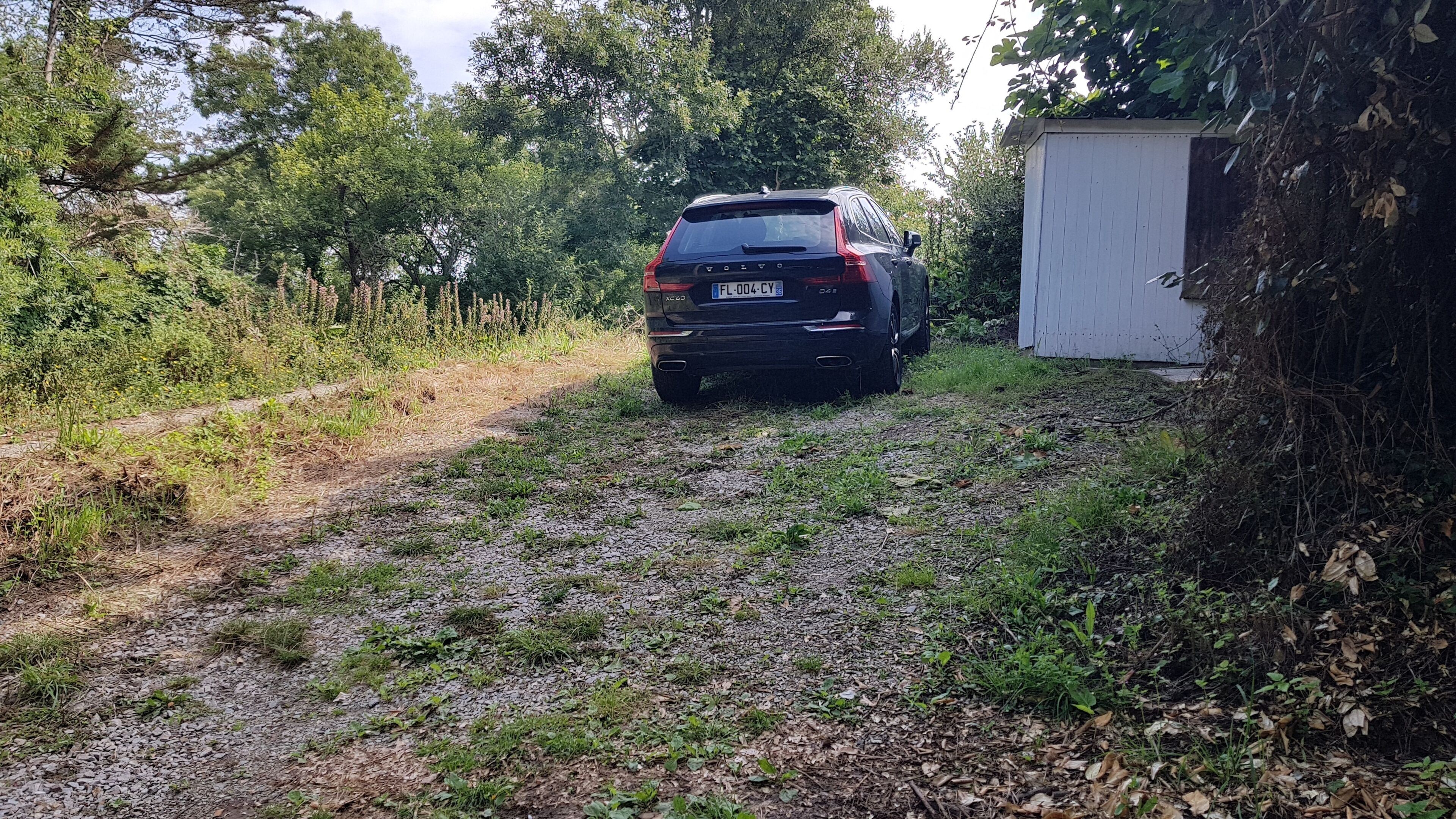 Une voiture noire était garée sur un chemin de gravier, à côté d’un abri blanc situé dans une zone boisée.