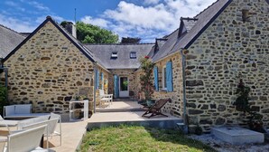 Un edificio de piedra con tejado de tejas, un pequeño jardín con mobiliario exterior y una entrada estrecha.