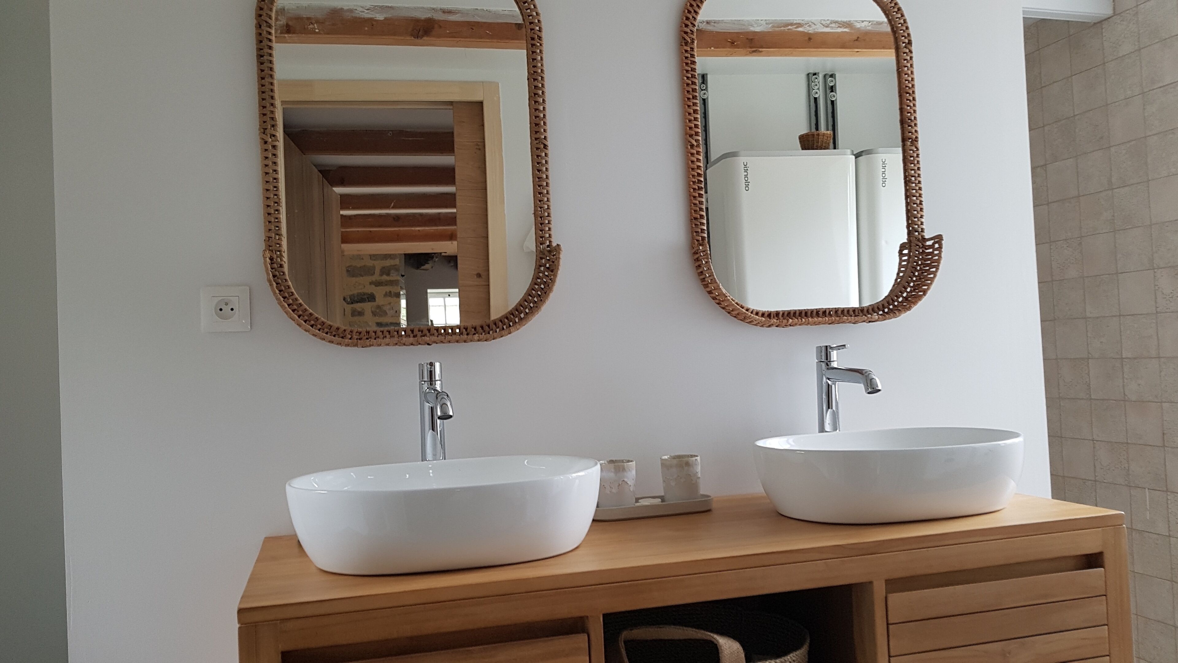 Une salle de bain avec deux vasques blanches, un meuble-lavabo en bois et deux grands miroirs dotés de cadres tressés.
