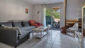 Een moderne woonkamer met een grijze bank, een salontafel en een witte stoel. Er is een schuifdeur naar een buitengebied met een tafel en stoelen.