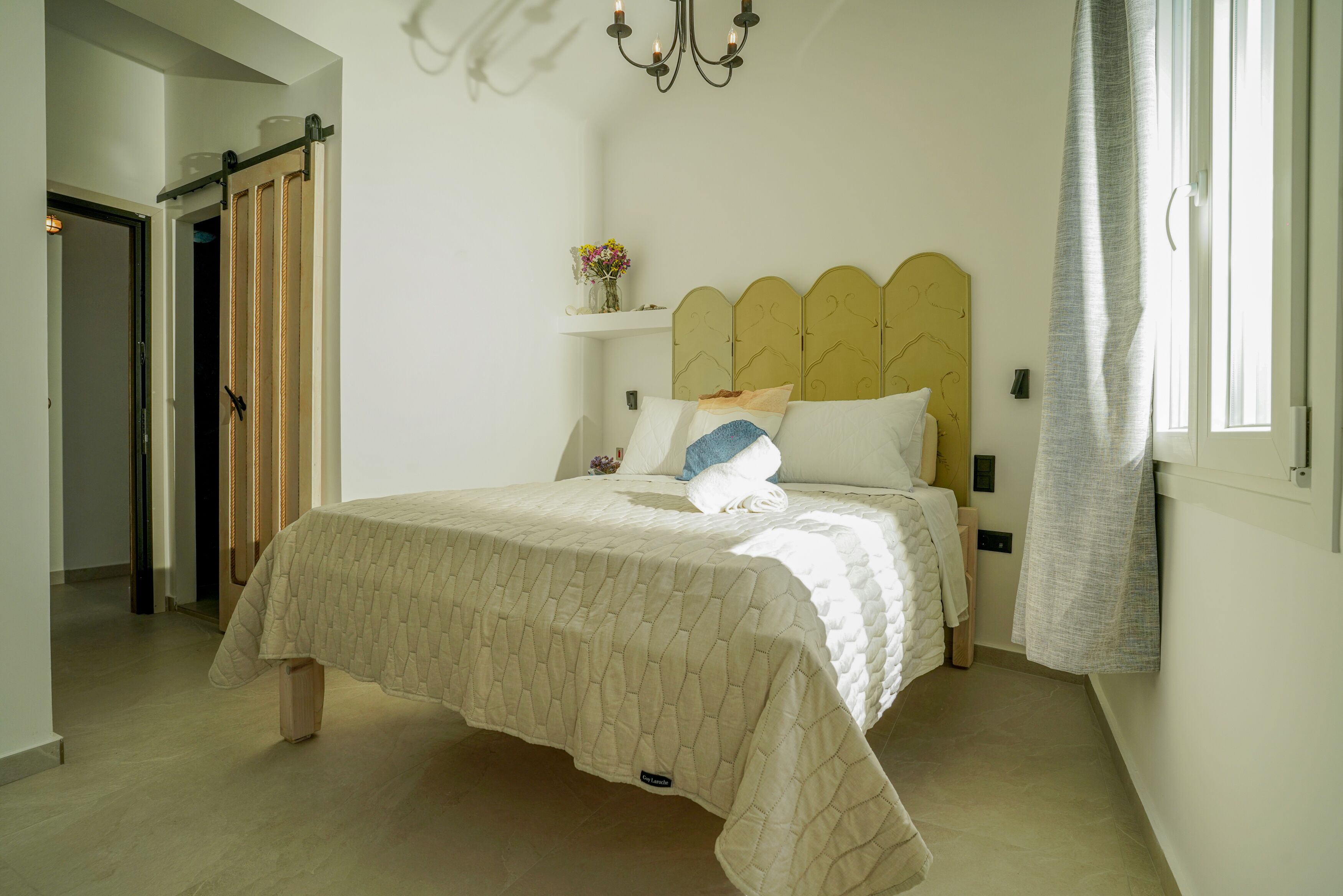 A bedroom with a bed, a chandelier, a window with curtains, and a door.