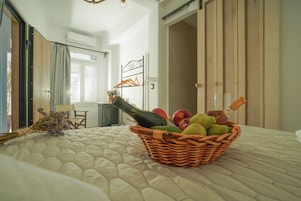 Um quarto com cama, uma cesta de vime com frutas e uma garrafa, uma janela e uma porta.