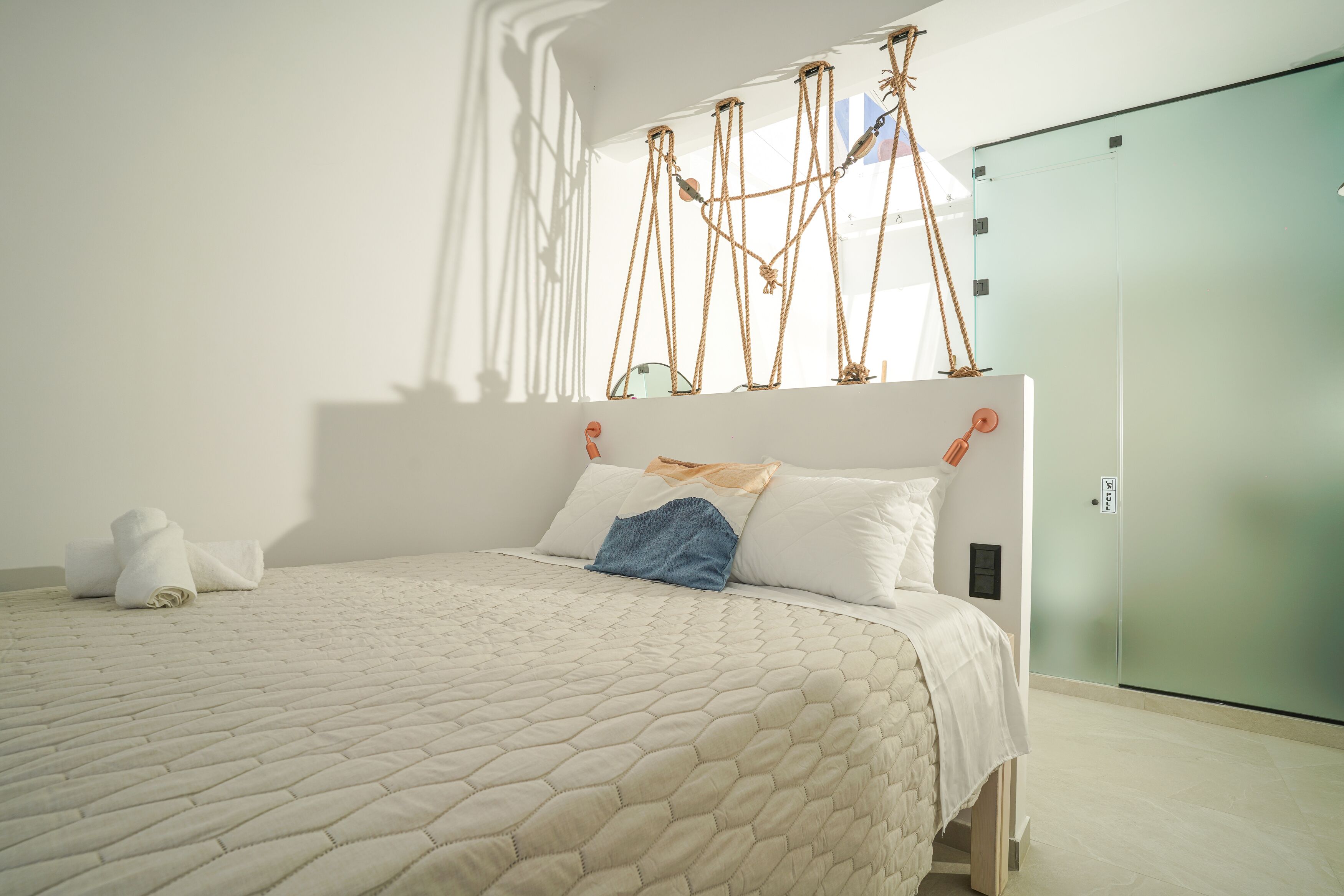 A neatly made bed with a quilted cover, a blue pillow, and a rolled towel on the left side.