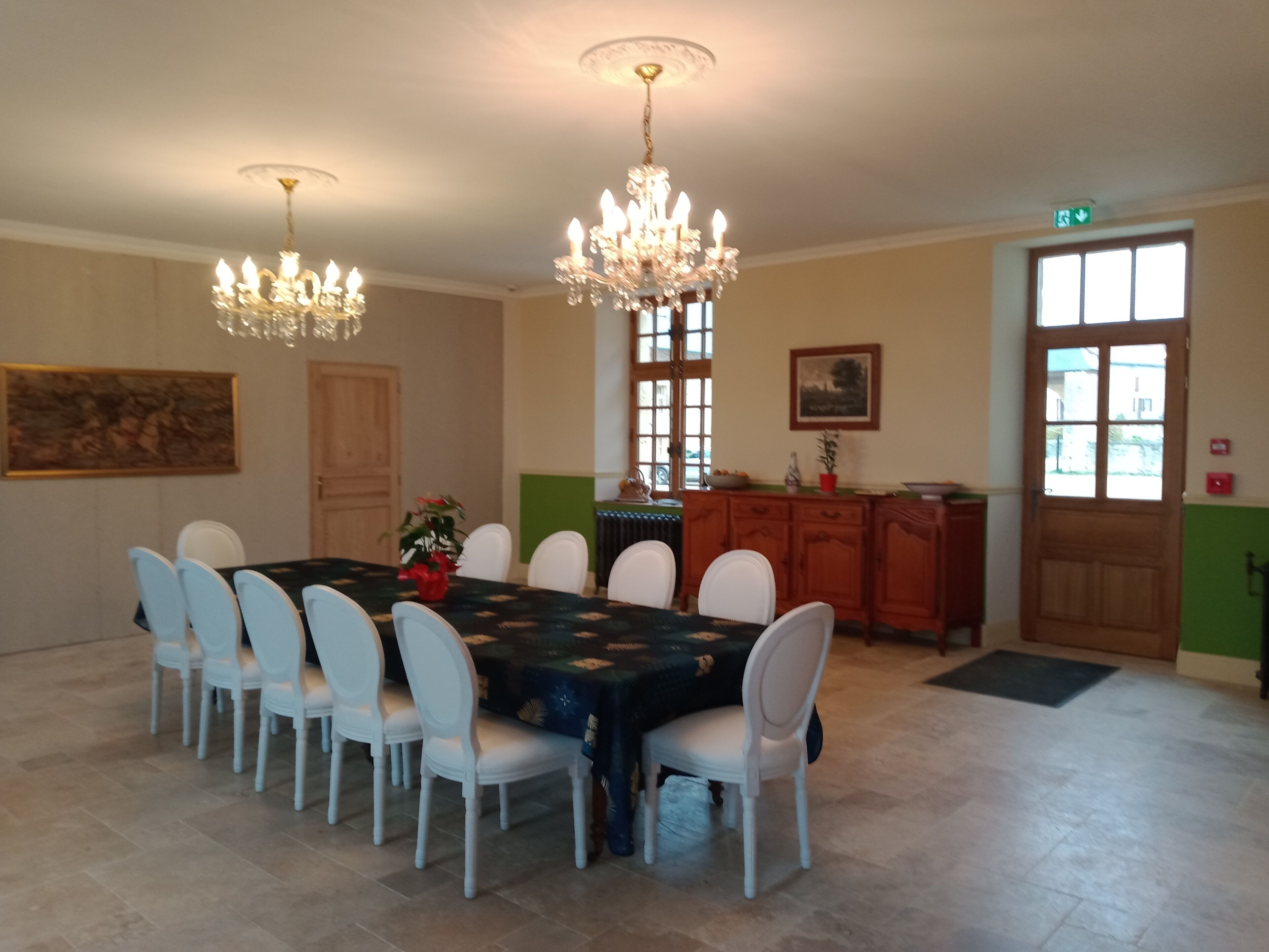 Un coin repas avec une table dressée pour six personnes, entourée de chaises blanches. Deux lustres pendent du plafond, ainsi qu’un buffet en bois avec un panneau vert et une porte vitrée.