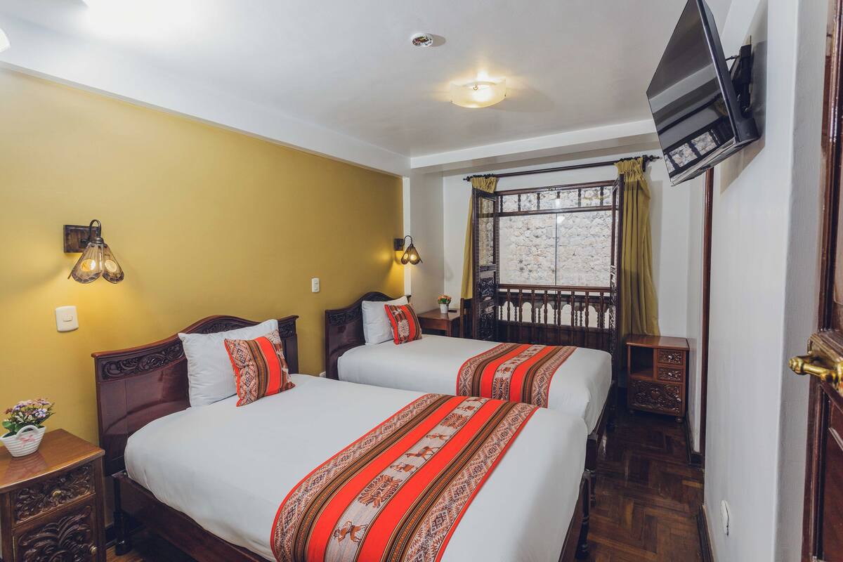 A hotel room with two beds, a TV mounted on the wall, a window with curtains, and a small table with a plant.