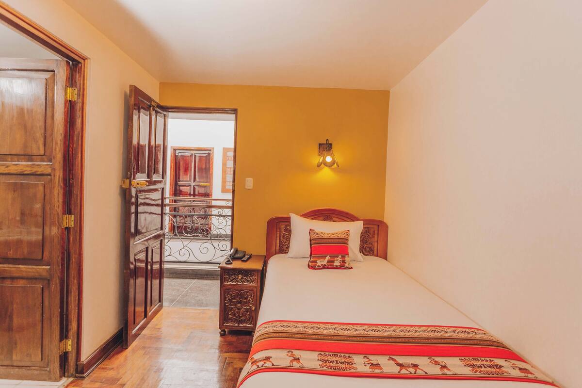 A bedroom with a wooden wardrobe, a bed with patterned bedding, a nightstand, and a wall-mounted light fixture.