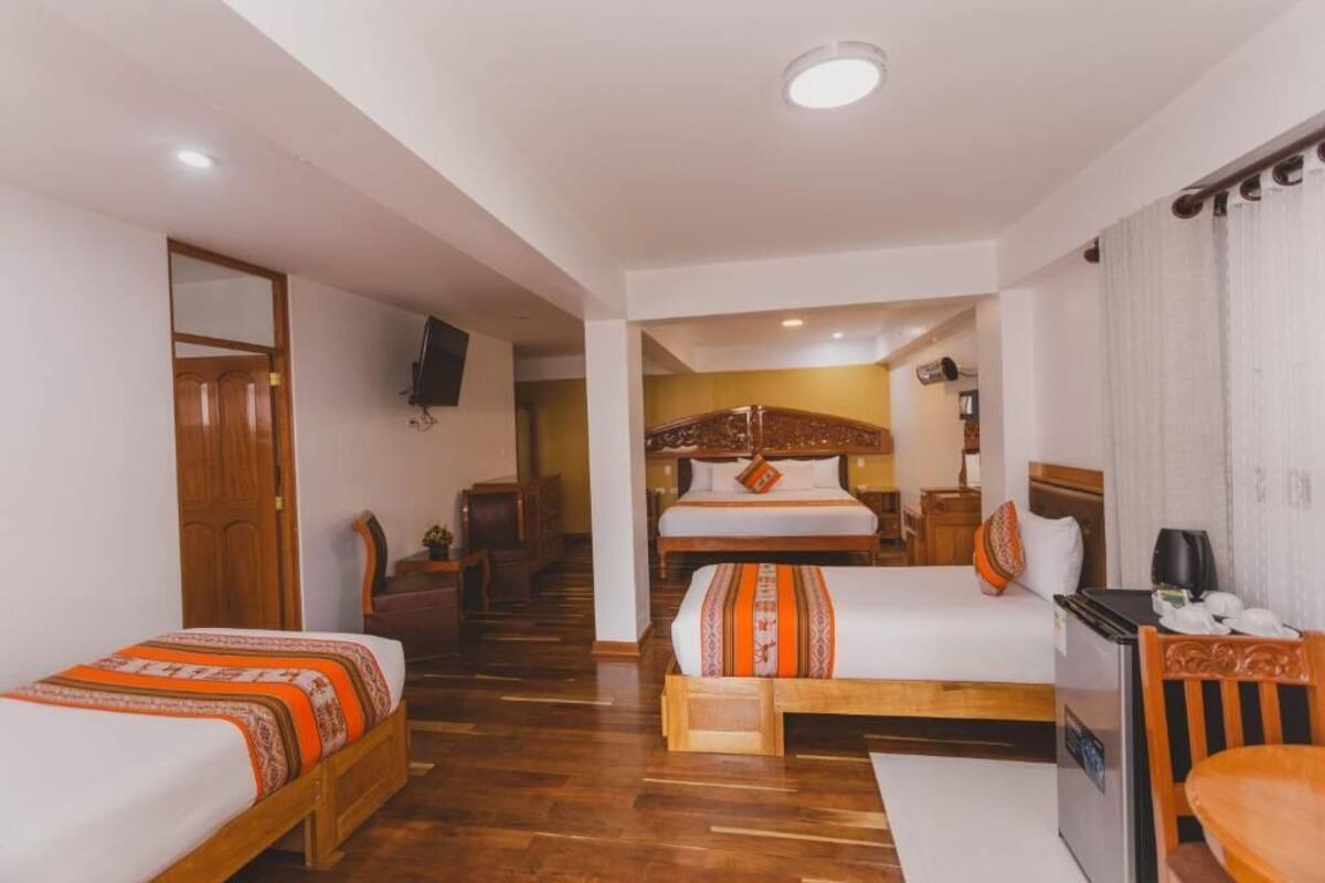 A hotel room with a wooden floor, a bed with a patterned bedspread, a desk with a chair, a TV mounted on the wall, and a door leading to another room.