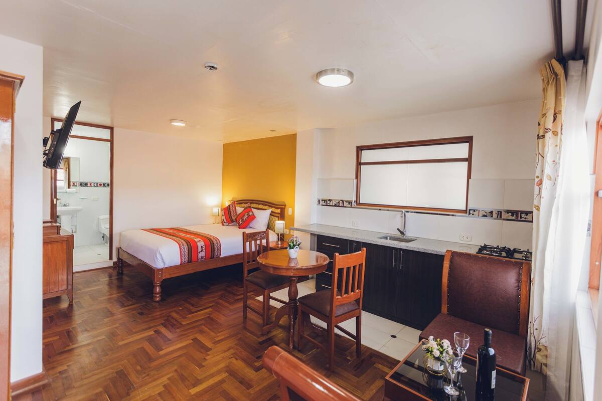 A compact hotel room with a bed, a small dining table, a chair, a sofa, a kitchen area, and a bathroom.