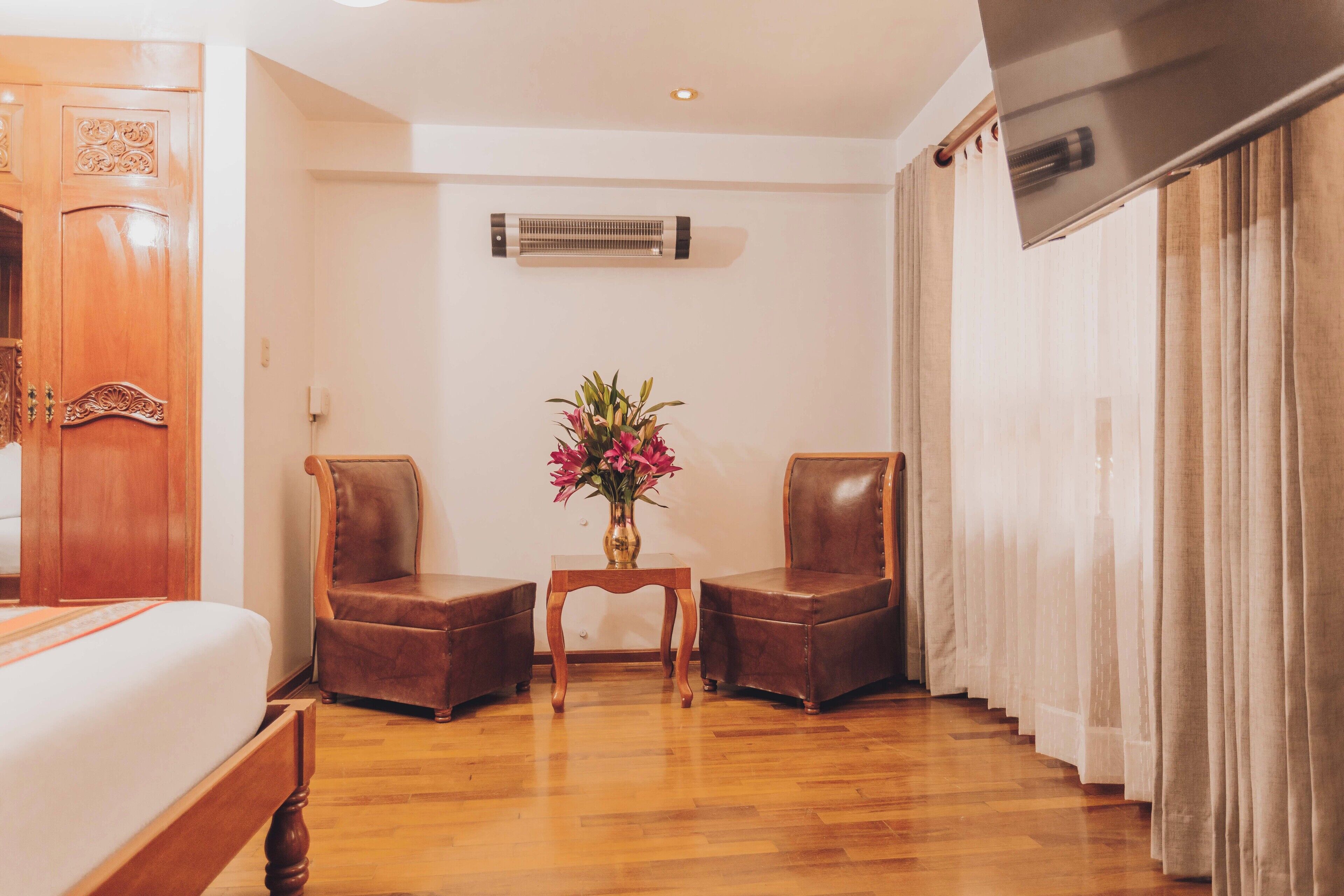 A hotel room with a wooden bed, two leather armchairs, a small wooden table with a vase of flowers, an air conditioning unit, and a large window with curtains.