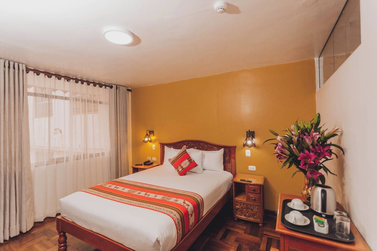 A hotel room with a bed, bedside tables, a nightstand, a vase with flowers, and a tray with cups and a kettle.