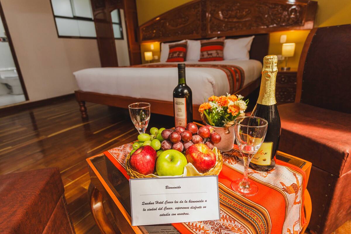 A hotel room with a bed, a wooden headboard, a nightstand with a lamp, and a table with a fruit basket, wine bottle, and glasses.